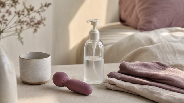 A compact vibrator, unscented lubricant and soft folded linen arranged in a calm bedroom setting