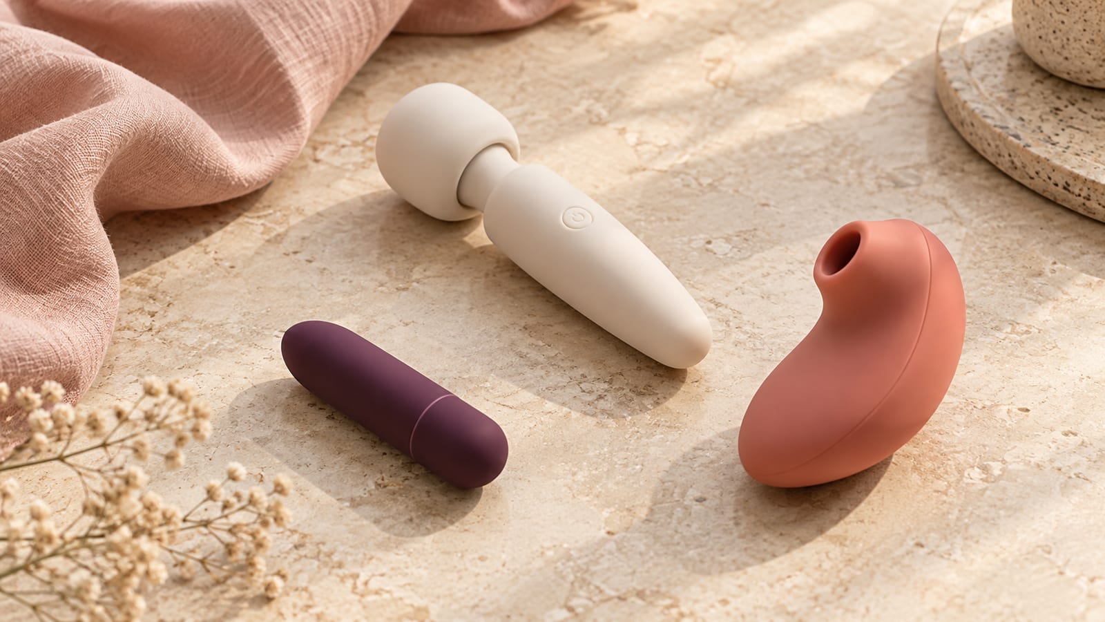 A plum bullet vibrator, ivory compact wand and coral air-pulse device arranged on warm stone beside blush linen.
